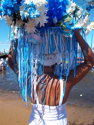 Lady holding large basket above head covered in beads and flowers, by the seaside