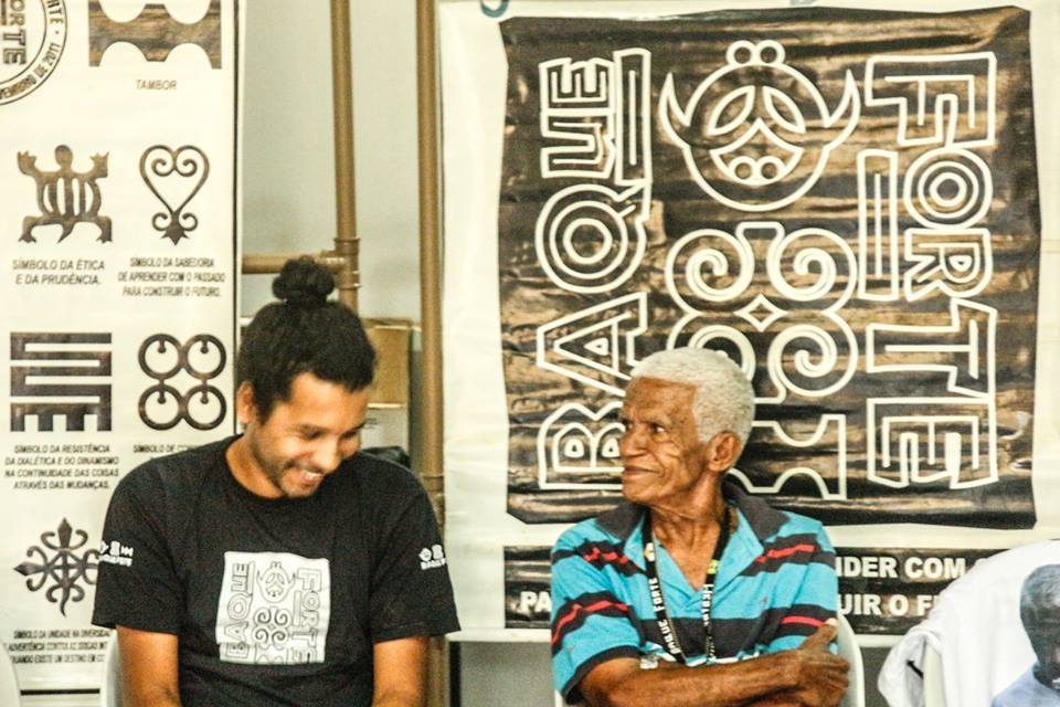 Mestre Jamesson and Mestre Toinho, sitting in front of a black and white flag, laughing