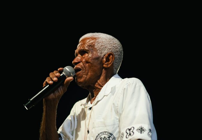 Mestre Toinho, holding a microphone and singing, wearing a white shirt against a dark background